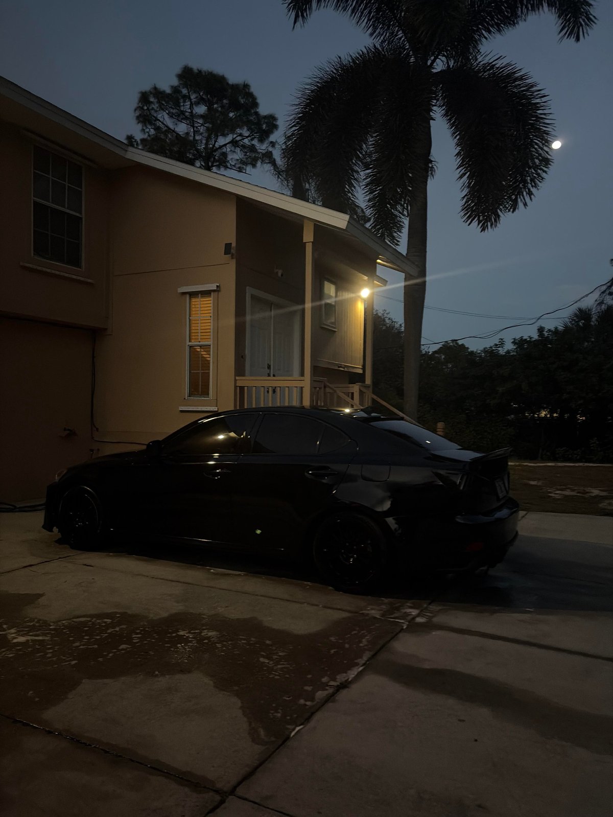 Black car parked in driveway of illuminated modern house at dusk with palm tree and trees in background
